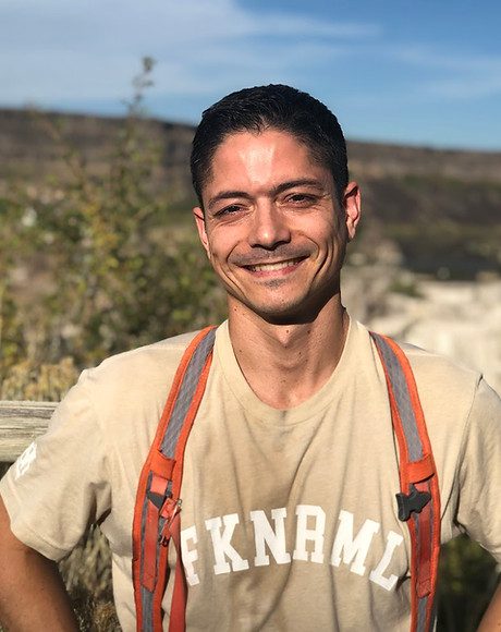 Eric on a trail above a high-desert canyon, in a sun-faded t-shirt and pack straps, smiling at the camera.